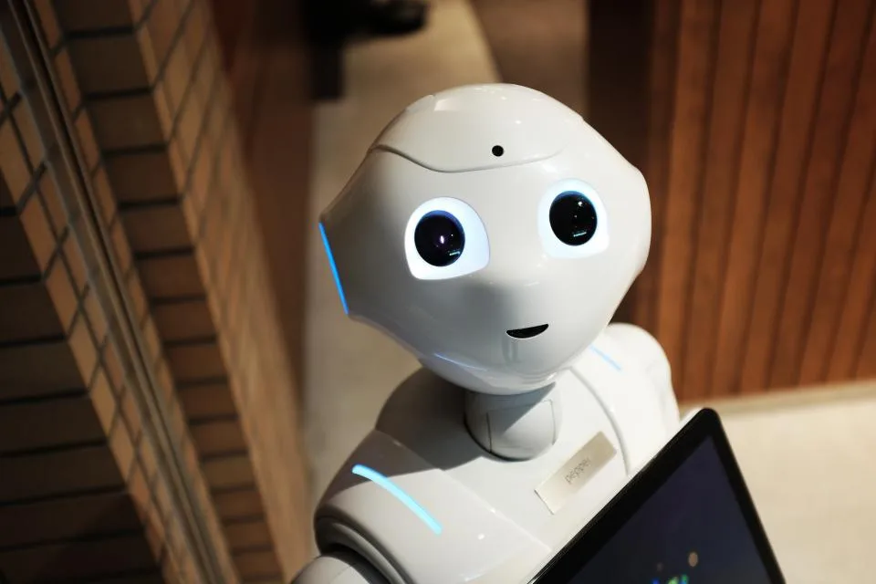 Robô de atendimento branco diante de um notebook, em ambiente de madeira