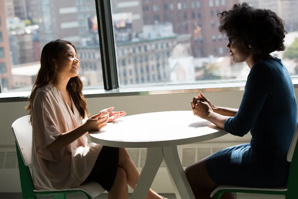 Duas profissionais conversando frente a frente a uma mesa, com a cidade ao fundo