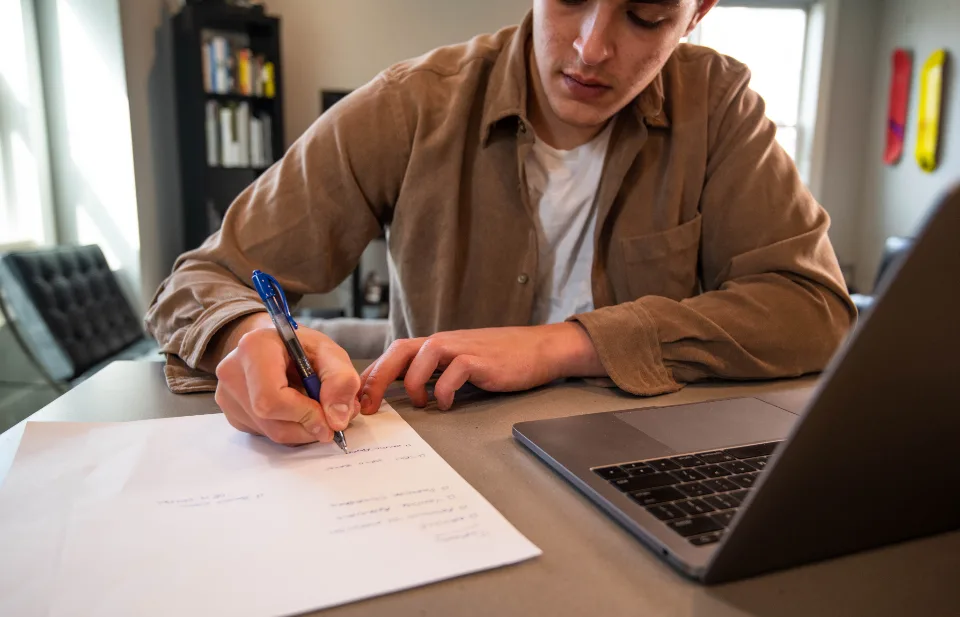 Homem escrevendo à mão numa folha ao lado do notebook